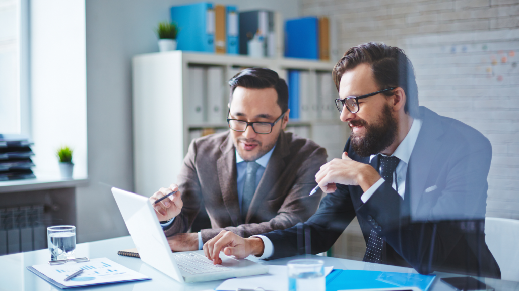 Two men with glasses and dressed in business suits look at a computer as if going over consulting